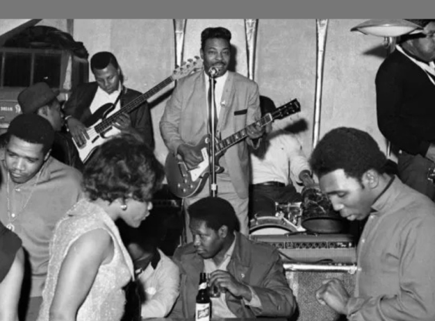 Black and white photo of a lively 1960s club scene. A band with a guitarist, bassist, drummer, and saxophonist plays on stage while people dance energetically.