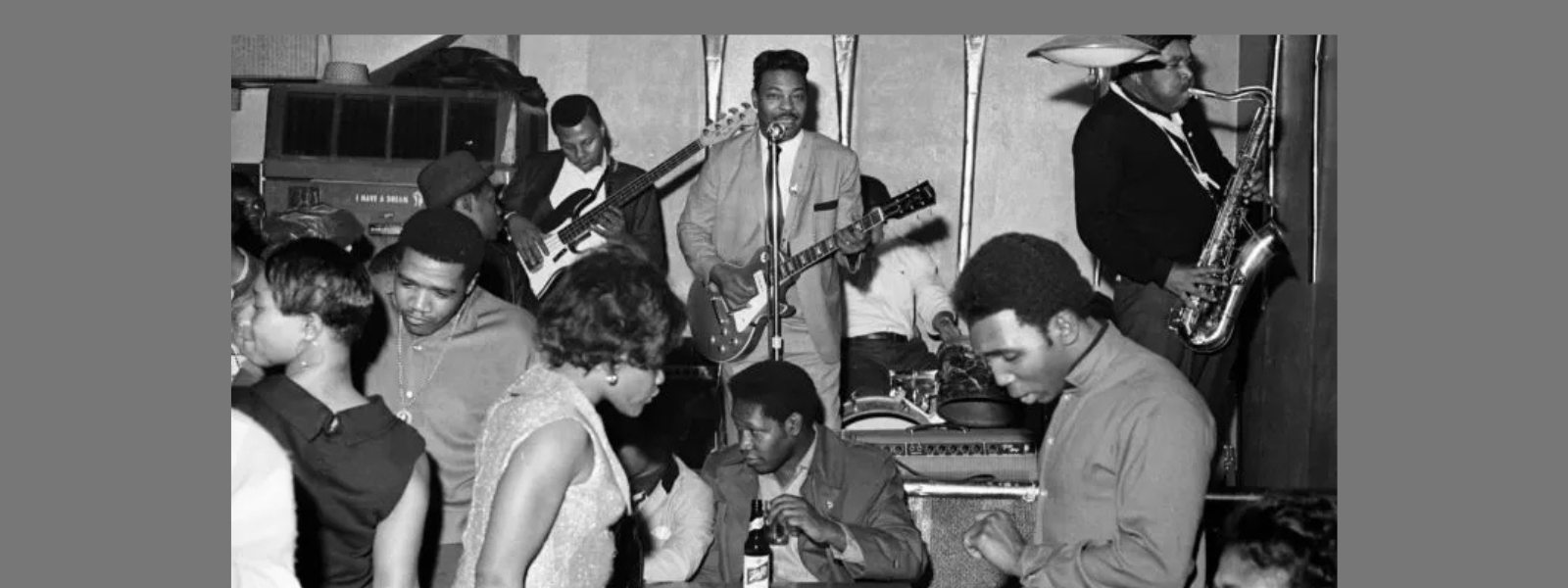 Black and white photo of a lively 1960s club scene. A band with a guitarist, bassist, drummer, and saxophonist plays on stage while people dance energetically.
