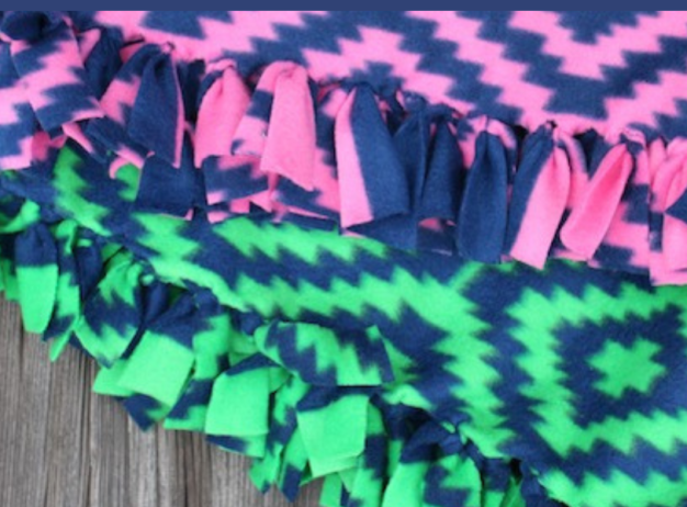 Two fleece blankets with zigzag patterns and knotted fringe edges. One is pink and navy, the other green and navy, placed on a wooden surface.