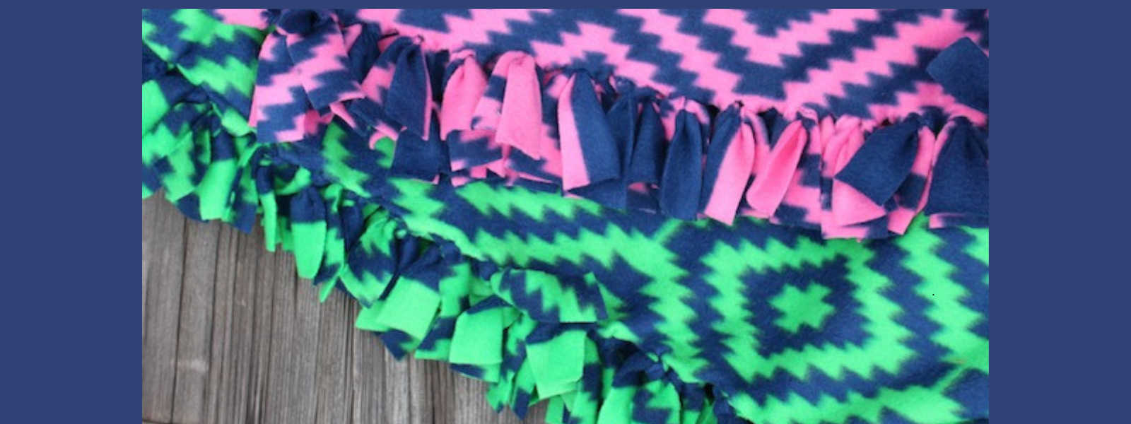 Two fleece blankets with zigzag patterns and knotted fringe edges. One is pink and navy, the other green and navy, placed on a wooden surface.