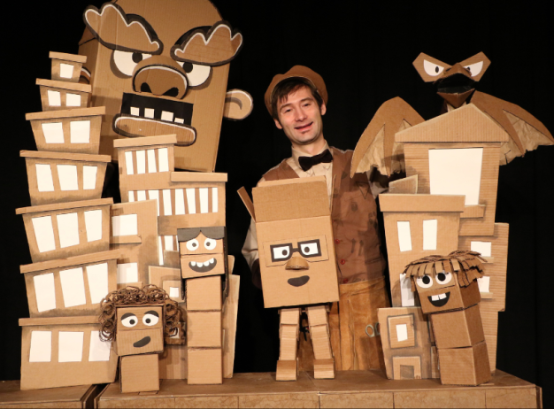 A performer stands with a playful expression surrounded by whimsical cardboard cityscape props and characters with expressive faces and features.