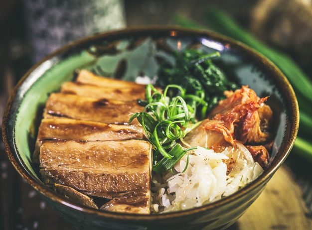 A bowl of rice topped with glazed, sliced pork, vibrant green spinach, kimchi, and sliced green onions, creating a fresh and flavorful presentation.