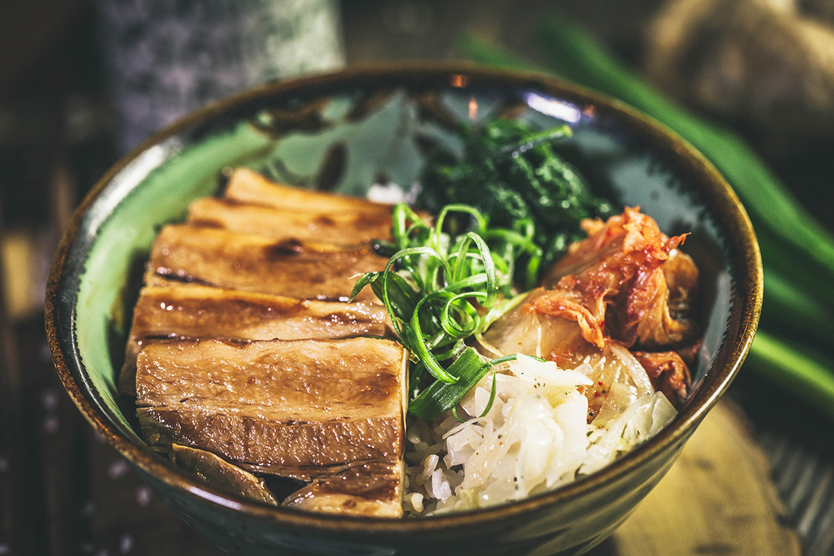 A bowl of rice topped with glazed, sliced pork, vibrant green spinach, kimchi, and sliced green onions, creating a fresh and flavorful presentation.