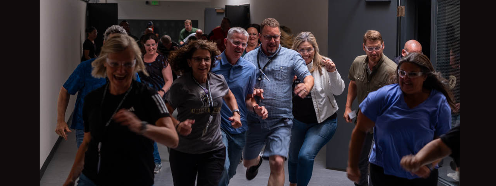 A group of people enthusiastically running down a hallway, smiling and dressed casually. The mood is energetic and lively, with bright overhead lights.