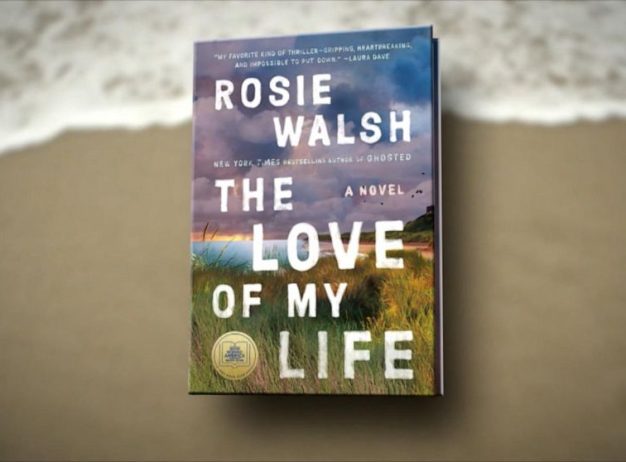 Book cover of "The Love of My Life" by Rosie Walsh on a sandy beach background. The cover depicts a coastal scene with grass and a purple sky.