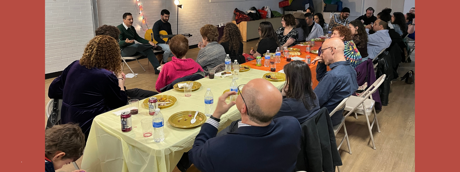 A group of people sit around tables with yellow and orange tablecloths, listening to two speakers. The mood is attentive and sociable.