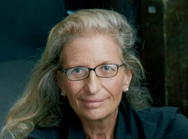 Portrait of an older individual with glasses and long, wavy gray hair, wearing a dark top. They have a calm expression, set against a soft-focus dark background.