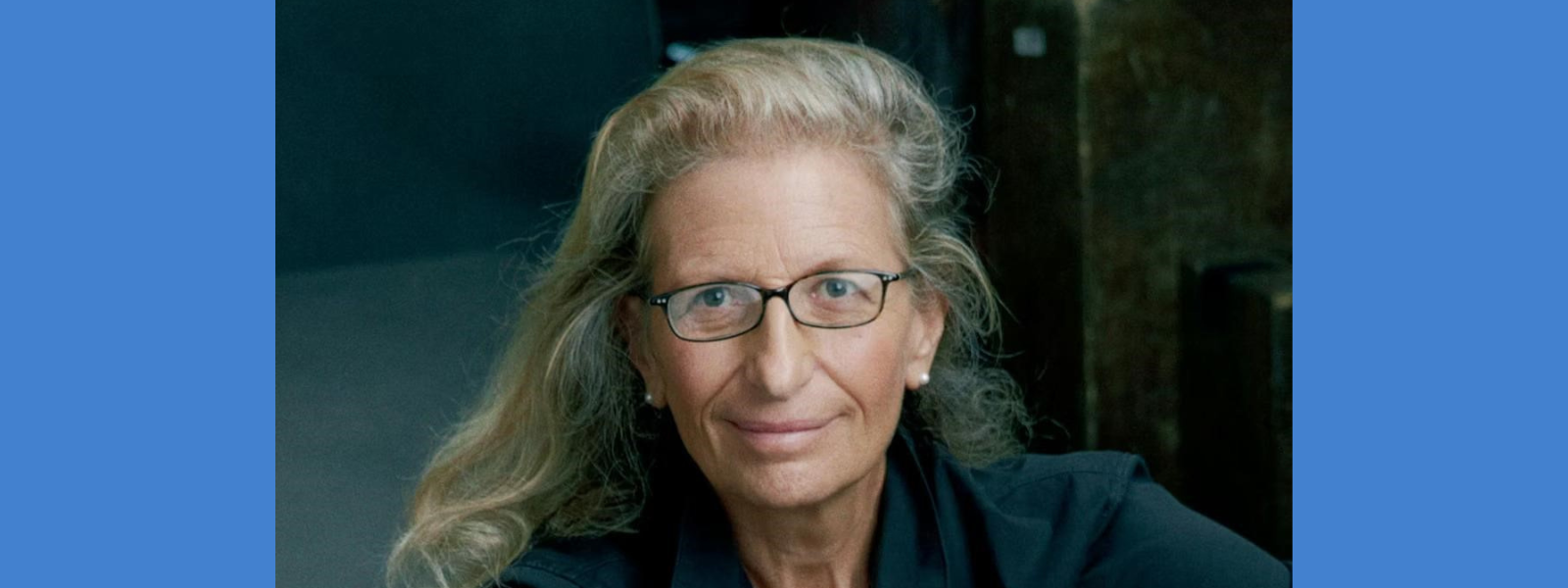 Portrait of an older individual with glasses and long, wavy gray hair, wearing a dark top. They have a calm expression, set against a soft-focus dark background.
