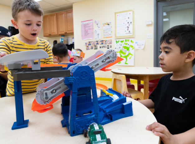 Two young boys are focused and engaged in playing with a blue and orange toy car track on a classroom table. Children in the background add to the lively atmosphere.