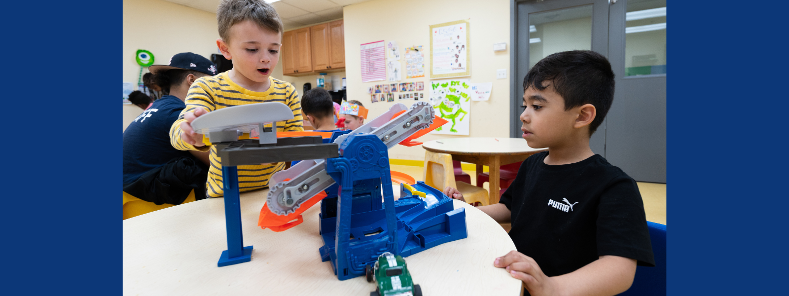 Two young boys are focused and engaged in playing with a blue and orange toy car track on a classroom table. Children in the background add to the lively atmosphere.