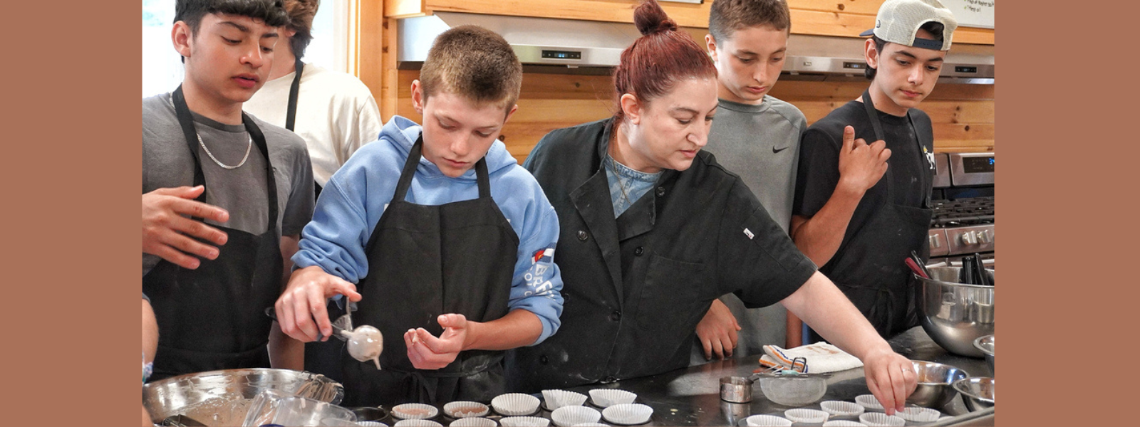 A group of teens and an instructor are baking in a kitchen. They are focused on filling muffin cups with batter. The atmosphere is collaborative and focused.