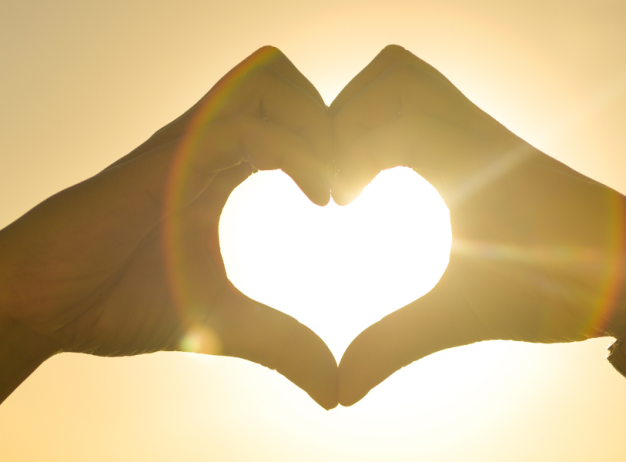 Hands forming a heart shape against a bright sunset, with sunlight streaming through. Warm, peaceful, and hopeful atmosphere.