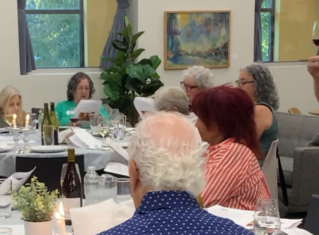 A group of people sit around a table in a well-lit room, enjoying a meal. One man stands, raising a glass of red wine, appearing to give a toast.