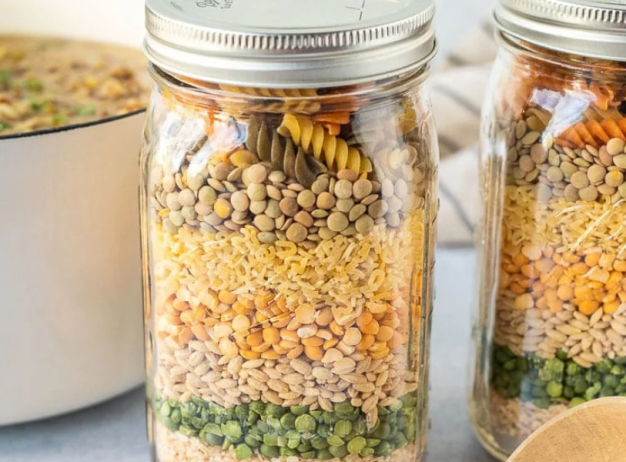 Two jars filled with layered pasta and dried lentils, beans, and peas sit on a countertop, with a pot of soup in the background, evoking a cozy kitchen feel.