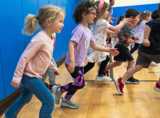 Children enthusiastically running in a gym, smiling and focused. The gym has blue walls, wooden floors, and set up cones in the background.