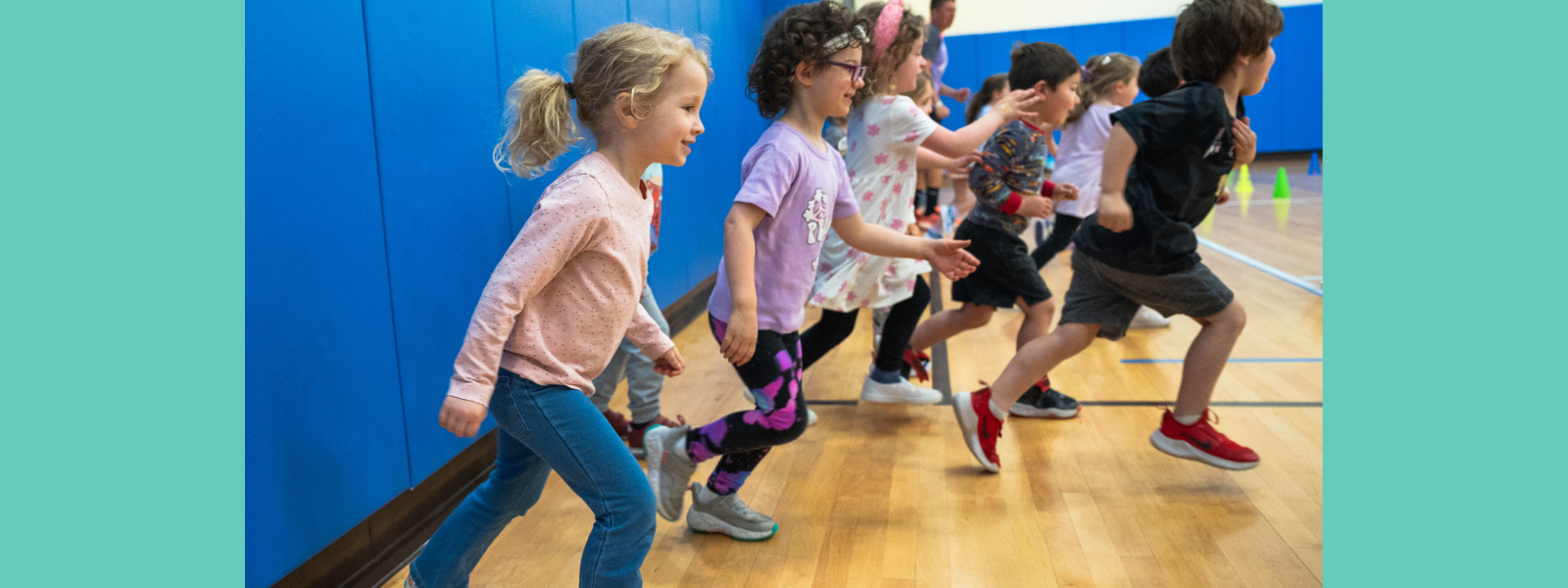 Children enthusiastically running in a gym, smiling and focused. The gym has blue walls, wooden floors, and set up cones in the background.