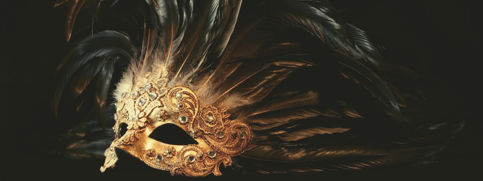 Ornate gold masquerade mask adorned with intricate patterns and dark feathers set against a black background, evoking a sense of mystery and elegance.