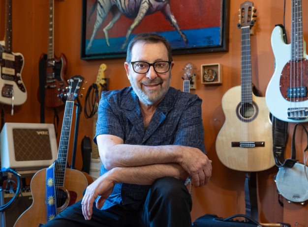 A smiling person with glasses sits in a room adorned with various guitars and a banjo, reflecting a warm, inviting, and musical atmosphere.