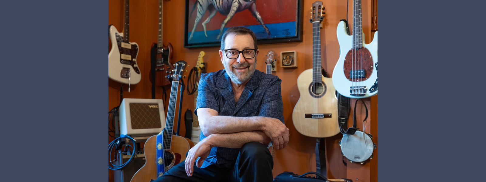 A smiling person with glasses sits in a room adorned with various guitars and a banjo, reflecting a warm, inviting, and musical atmosphere.