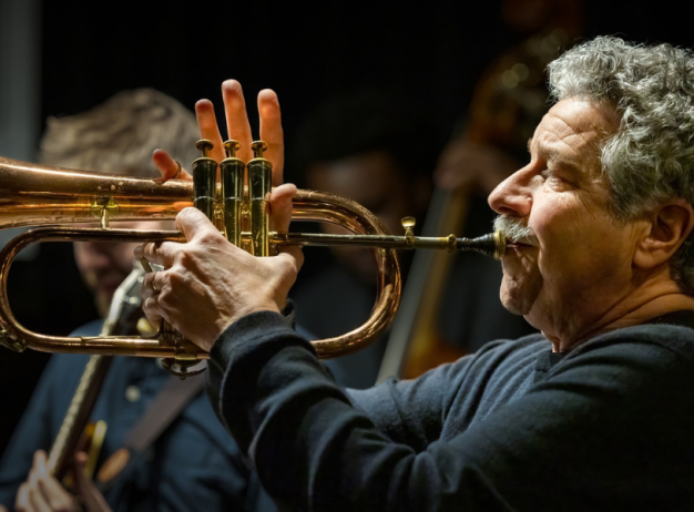 A man passionately plays a trumpet on stage, surrounded by blurred musicians. The warm lighting highlights his focused expression and the brass instrument.