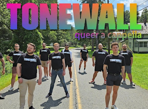 A group of people stand confidently on a rural road, wearing matching black "Tonewall" shirts. Above them, colorful "Tonewall" text reads "queer a cappella."