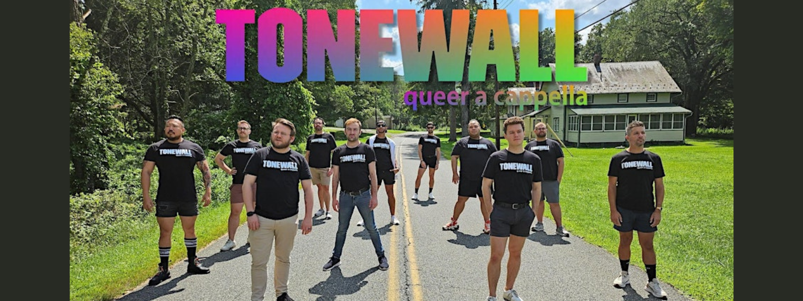A group of people stand confidently on a rural road, wearing matching black "Tonewall" shirts. Above them, colorful "Tonewall" text reads "queer a cappella."