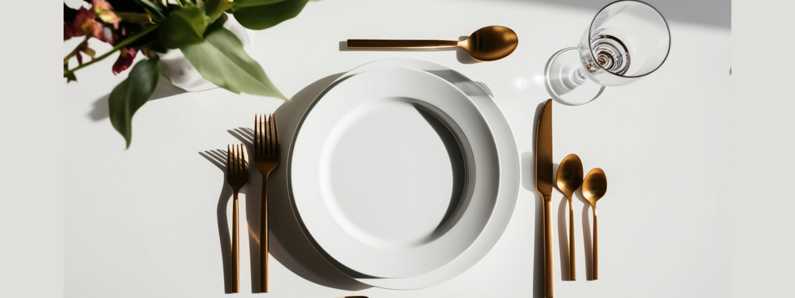 Elegant table setting with a white plate, gold utensils, and a wine glass on a white table. A plant with flowers adds a touch of green and warmth.