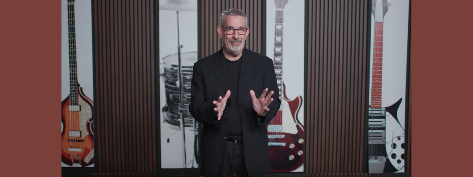 A smiling man in a black suit stands in front of a backdrop featuring large guitar images. The background suggests a musical context, setting a professional yet relaxed tone.
