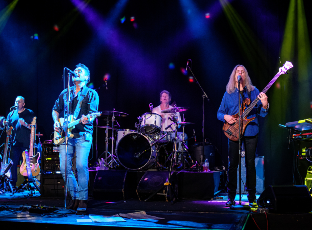 A six-member band performs on stage under vibrant blue, purple, and green lights. Musicians play guitar, drums, and keyboard, creating an energetic atmosphere.