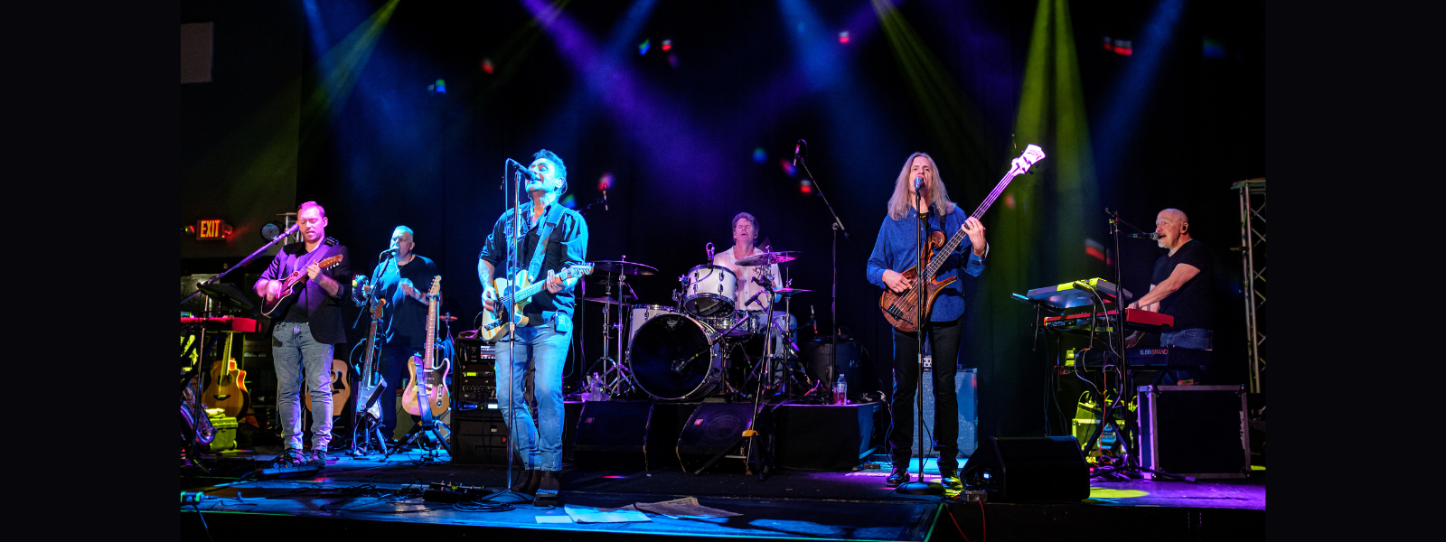 A six-member band performs on stage under vibrant blue, purple, and green lights. Musicians play guitar, drums, and keyboard, creating an energetic atmosphere.